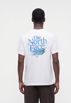 Mies, jolla on päällä valkoinen T-paita, jossa on sininen ja vihreä "The North Face" logo ja kuva selässä, yhdistetty tummanruskeisiin housuihin.