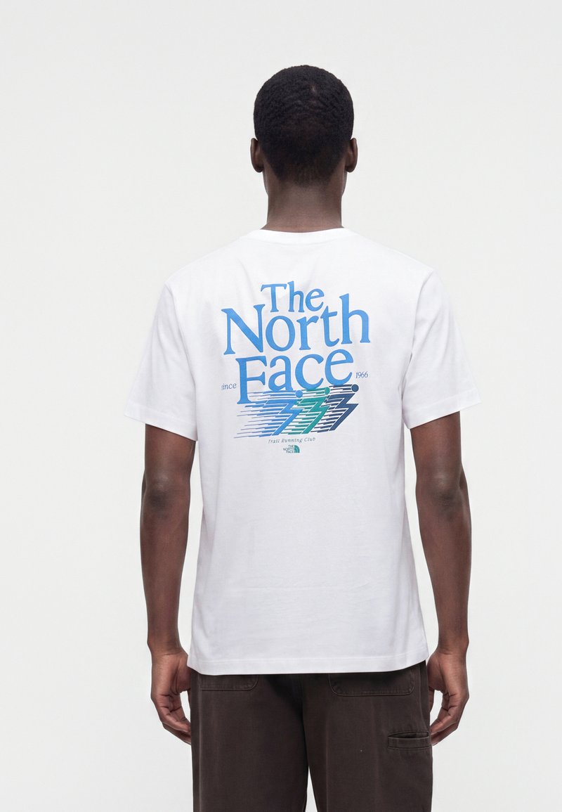 Άνδρας που φοράει λευκό T-shirt με το λογότυπο και το γραφικό "The North Face" σε μπλε και πράσινο στο πίσω μέρος, συνδυασμένο με σκούρο καφέ παντελόνι.