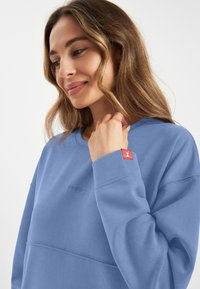 Lichtblauwe sweatshirt met een ronde hals, ribgebreide manchetten en geborduurd logo. Heeft een klein rood accentlabel op de mouw. Zachte textuur.