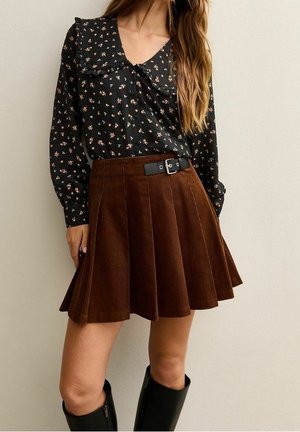 Vrouw draagt een zwarte bloes met lange mouwen met bloemenprint, ingestopt in een bruine geplooide corduroy rok met een zwarte riem en zwarte kniehoge laarzen.
