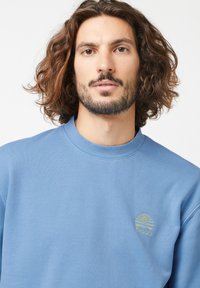 Blauwe crewneck sweatshirt van zachte stof, met een klein groen geborduurd logo op de linkerborst en een relaxte pasvorm.