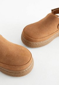 Sabots en daim marron avec un bout arrondi, une semelle texturée et des détails couturés. Le design présente un dos ouvert et une semelle intérieure rembourrée.