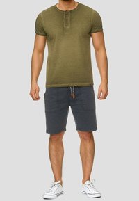 T-shirt henley manches courtes couleur vert olive avec trois boutons ; associé à un short gris foncé avec deux poches latérales et un cordon de serrage. Baskets blanches.