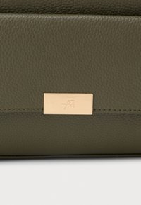 Borsa in pelle verde oliva con superficie texturizzata, dotata di un'etichetta in metallo color oro. Linee pulite e forma rettangolare compatta.