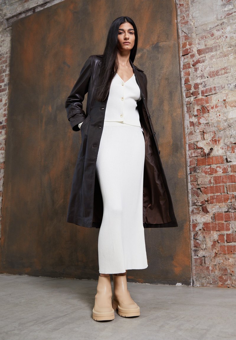 Casaco trench de couro castanho sobre um vestido branco com texturas em nervuras, apresentando um decote em V profundo e detalhes em botões; combinado com botas de plataforma bege.