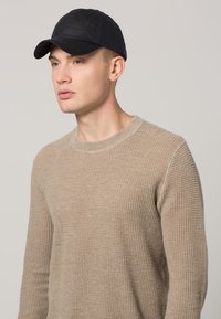 Pulover tricotat texturat bej cu guler rotund, asortat cu o șapcă de baseball neagră. Materialul pare moale, cu o ușoară elasticitate.
