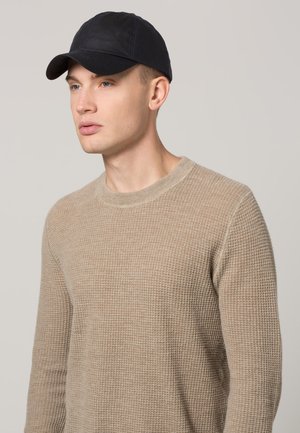 Beige pletený svetr s texturou a kulatým výstřihem, doplněný černou baseballovou čepicí. Tkanina vypadá měkce s mírným strečem.