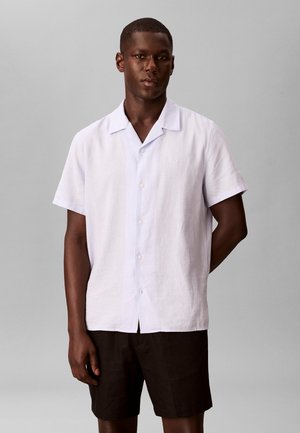 Uomo in piedi che indossa una camicia bianca a maniche corte con bottoni e pantaloncini scuri davanti a uno sfondo grigio uniforme.