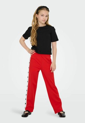 Zwarte T-shirt met korte mouwen gecombineerd met rode wijde broek met zwarte stippen aan de zijkant. Gdragen met zwarte schoenen.