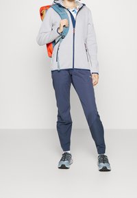 Gray fleece jacket with blue accents, zippered front, paired with navy pants and light blue hiking shoes. Model carrying an orange backpack.