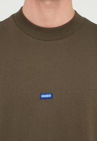 Olivegrønn bomulls-T-skjorte med rund hals, med en blå rektangulær "HUGO" logo på forsiden, med ribbestrikket halskant og jevn tekstur.