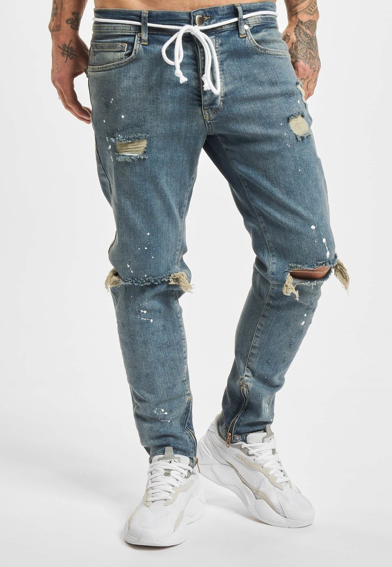 Onderlichaam met gescheurde blauwe spijkerbroek met wit trekkoord en witte sneakers, zichtbare getatoeëerde armen aan de zijkanten.