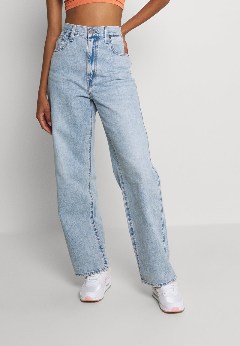 Personne portant un jean taille haute bleu clair à jambes larges et des baskets blanches, debout avec une main dans la poche sur un fond uni.