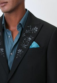 Schwarzer Blazer mit floraler Bestickung am Revers, kombiniert mit einem türkisfarbenen Hemd und einem türkisfarbenen Einstecktuch, aus strukturiertem Stoff.