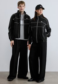 Μαύρα αδιάβροχα με λευκές λεπτομέρειες, φορεμένα πάνω από γκρι πουκάμισα, συνδυασμένα με φαρδιά μαύρα παντελόνια και μαύρα παπούτσια. Ένα μαύρο καπέλο ολοκληρώνει την εμφάνιση.