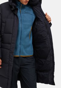 Person som bär en blå fleecejacka och svarta byxor, håller en lång svart vadderad kappa öppen för att visa sin innervinkel.
