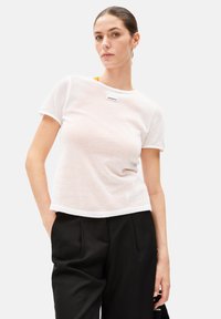 White mesh t-shirt with short sleeves, a round neckline, and a brand label on the chest. Paired with black high-waisted trousers.
