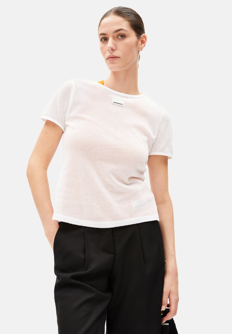 White mesh t-shirt with short sleeves, a round neckline, and a brand label on the chest. Paired with black high-waisted trousers.