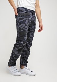 Man med svarta och grå camouflagetäckta cargobyxor, vit t-shirt och vita sneakers, stående med en hand i fickan.