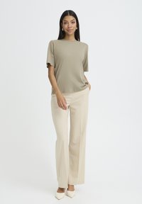 Kvinde står vendt mod fronten iført beige top med korte ærmer, cremefarvede bukser med vide ben, hvide spidse sko og sølvfarvede hoop øreringe.