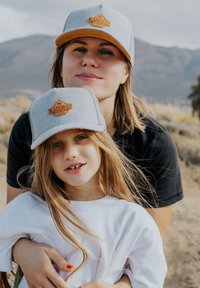 Zwei Personen tragen graue und beige Snapback-Mützen mit einem Lederpatch, das mit "BLACKSKIES" beschriftet ist. Der Hintergrund zeigt trockenes, grasiges Gelände.