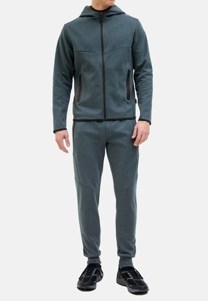 Felpa grigia con zip e cappuccio, dotata di due tasche laterali, abbinata a pantaloni della tuta a tapered, caratterizzati da una texture morbida e un design aderente.