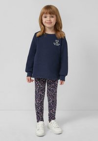 Granatowy sweter z białym graficznym sercem i napisem; legginsy w kwiatowy wzór w ciemnym odcieniu; białe sneakersy; dziewczyna z włosami do ramion.