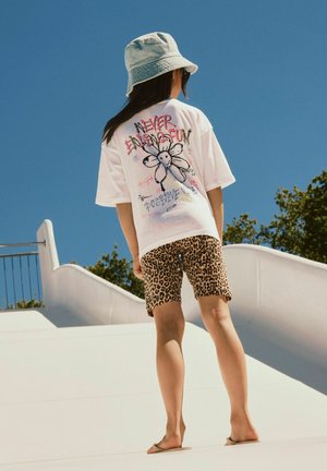 Enfant portant un bob, un t-shirt blanc à motifs avec une fleur et du texte, un short imprimé léopard et des sandales marchant pieds nus sur une surface blanche courbée.