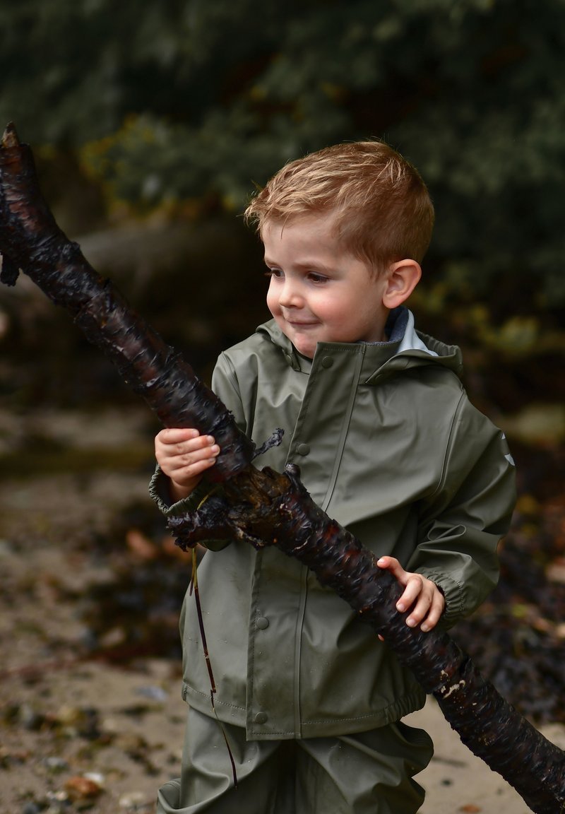 Un niño con una chaqueta impermeable verde sostiene un palo grande, oscuro y texturizado. La chaqueta tiene una cremallera y broches, con capucha.