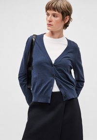Marineblauwe cardigan met een V-hals, vijf knopen aan de voorkant, een gebreide structuur en een geribbelde zoom. Gedragen over een wit shirt en gecombineerd met een zwarte rok.