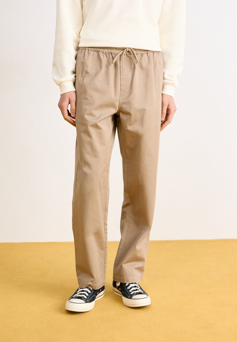 Bežové bavlnené jogger nohavice s elastickým pásom a šnúrkou. Skombinované s čiernymi plátenými teniskami s bielou gumovou podrážkou.