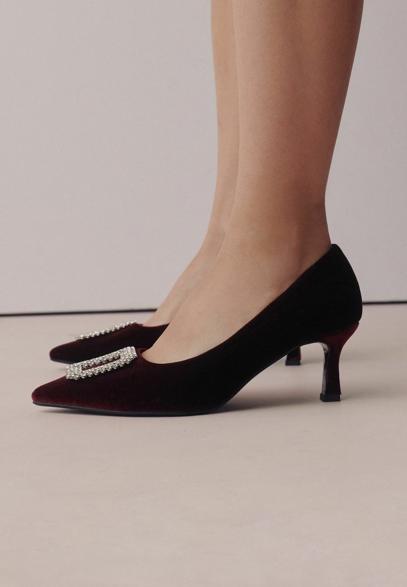 Pies con zapatos de tacón bajo de terciopelo rojo oscuro, con punteras puntiagudas y adornos rectangulares de piedras brillantes, sobre un fondo claro y liso.