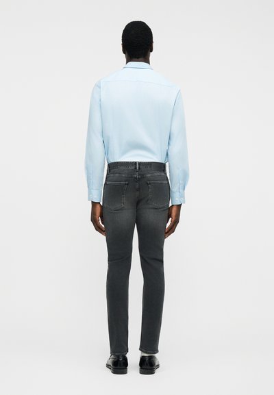 Άτομο που φοράει ανοιχτό μπλε πουκάμισο με μακριά μανίκια, σκούρο γκρι τζιν και μαύρα παπούτσια, στέκεται γυρισμένο με την πλάτη προς τα εμάς σε απλό φόντο.