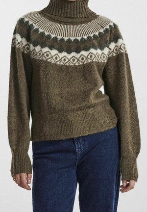 Femme portant un pull en laine marron à col roulé avec un empiècement blanc et marron foncé à motifs et un jean en denim bleu.