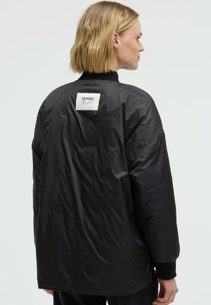 Black, padded jacket featuring a high collar, elastic cuffs, and a logo tag on the back. The material has a smooth, shiny texture.