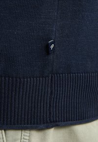 Marinblått stickat tröja med ribbat nedre kant, med en liten tygetikett på sidan som visar ett vitt bokstav "P". Slät textur.