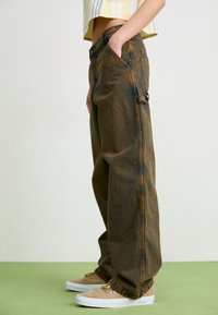 Jeans di denim marroni larghi con una trama sbiadita, dotati di tasche laterali e vita arricciata. Indossati con scarpe beige su una superficie verde.