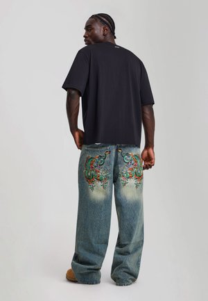 Mann mit geflochtenen Haaren, der ein schwarzes T-Shirt und weit geschnittene Jeans mit bunten bestickten Drachendesigns auf den Gesäßtaschen trägt.