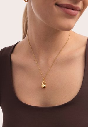 Collar de tono dorado con un pequeño colgante de candado. La cadena es delicada y retorcida, complementando una camiseta ajustada marrón.