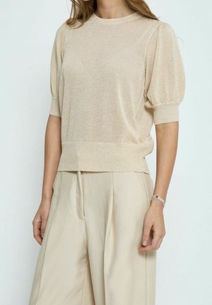 Femme portant un haut en maille beige à manches courtes bouffantes et un pantalon ample beige taille haute, debout devant un fond uni.