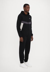 Black zip-up hoodie with a grey and white panel across the chest, paired with black sweatpants and white sneakers. Smooth fabric texture.
