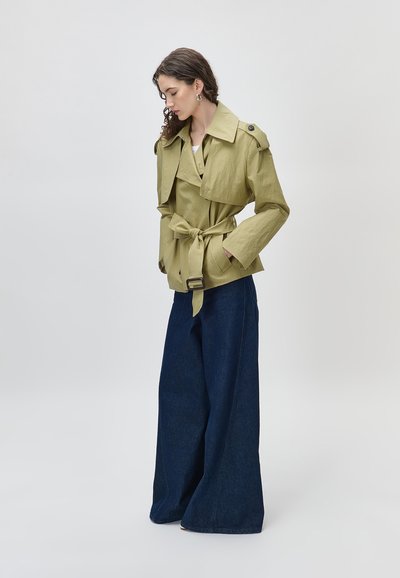 Γυναίκα που φοράει ανοιχτοπράσινο σακάκι με ζώνη και φαρδιές πέτες, καθώς και υπερμεγέθη ναυτικά μπλε παντελόνια με φαρδύ πόδι, στέκεται μπροστά σε απλό φόντο.