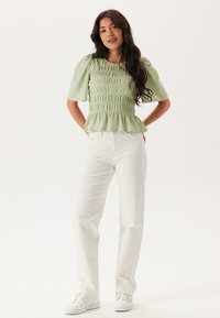 Blusa smock leggera verde chiaro con maniche corte a palloncino, abbinata a jeans bianchi a gamba dritta e sneakers bianche. I dettagli delle cuciture sono visibili.