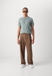 Lys grønn polo-skjorte med krage og tre knapper, kombinert med brune vide bukser og hvite joggesko. Jevn tekstur, avslappet passform.