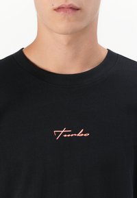 Close-up of a person wearing a black crewneck shirt with the word "Turbo" in small red cursive text at chest level.