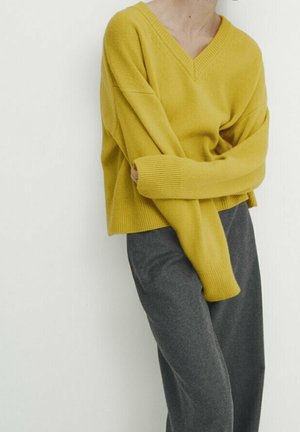Osoba ubrana w musztardowożółty sweter z dekoltem w kształcie litery V i długimi rękawami oraz ciemnoszare spodnie, stojąca na tle jednolitej białej ściany.