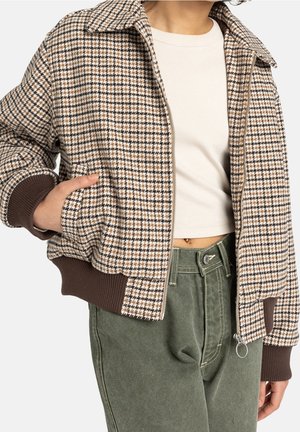 Person trägt braun-weißes Hahnentritt-Reißverschlussjacke, weißes kurzes Oberteil und hochtaillierte grüne Hose, eine Hand in der Tasche.