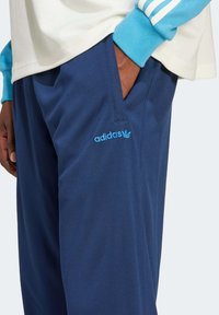 Navy blue athletic pants made of textured fabric, featuring an Adidas logo in light blue and side pockets for storage.