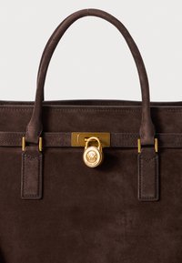 Bruin suède handtas met gouden accenten, voorzien van een slotdetail en symmetrische handvatten. Twee zijklauwen zijn bevestigd aan de tas.