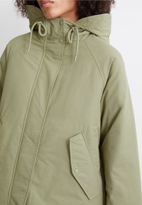 Khaki-grön jacka med hög krage, dragsko-huva och ficka med tryckknapp. Har ett mjukt, lätt tyg och raglanärmar.